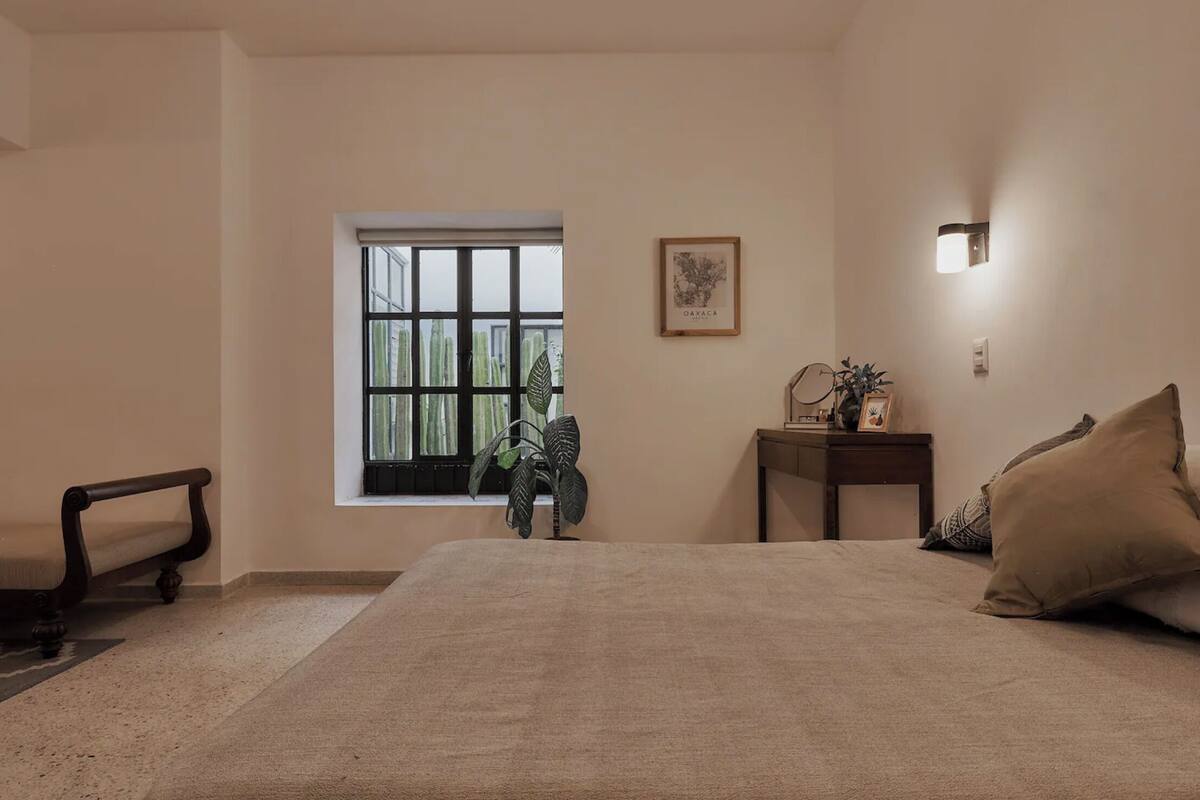 A bedroom with a large bed, a window with a view of trees, a bedside table with a lamp, and a wall-mounted light fixture.