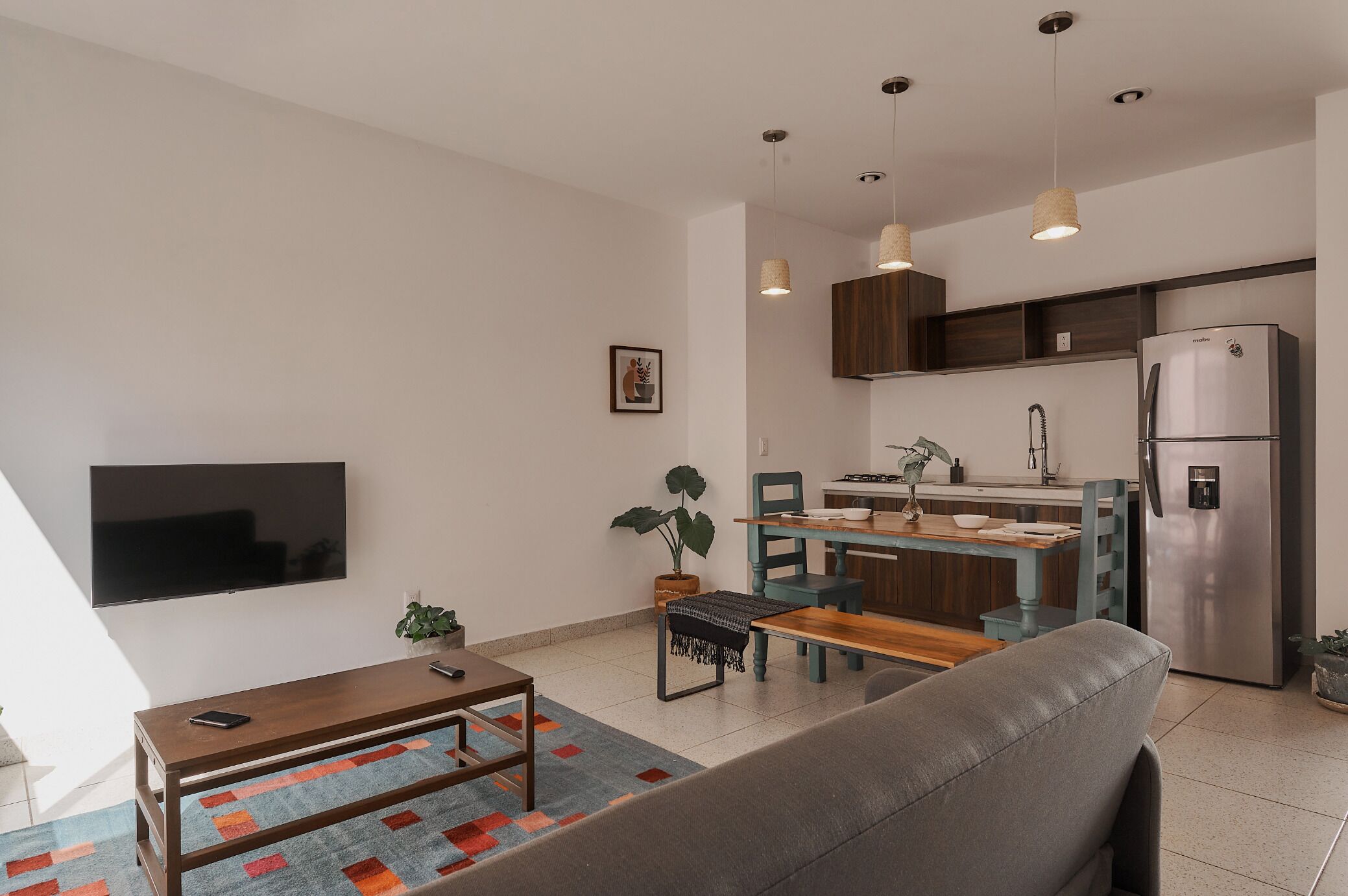 A modern living room with a flat-screen TV, a wooden coffee table, and a grey sofa. There is a kitchen area with a stainless steel refrigerator, a sink, and wooden cabinets.