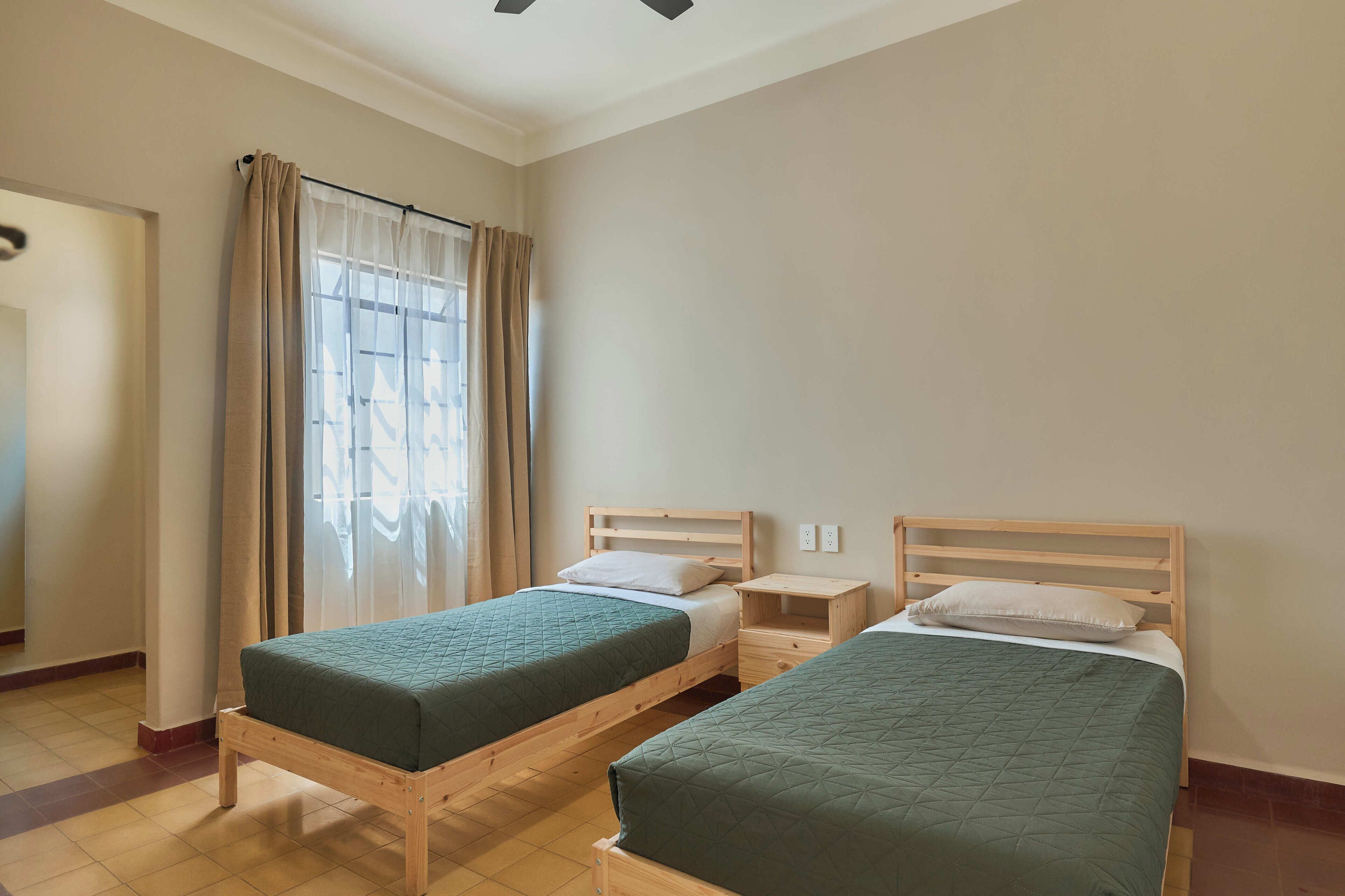 A room with two wooden beds, a window with curtains, and a small bedside table with a lamp.