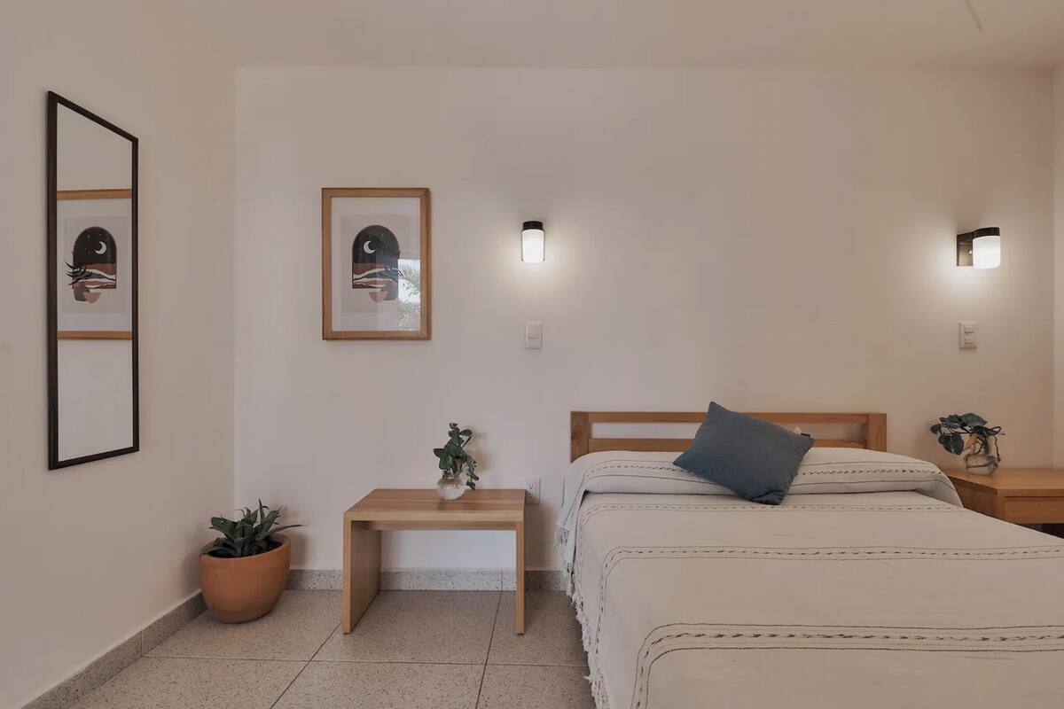 A bedroom with a bed, bedside tables, a nightstand, a potted plant, and wall-mounted light fixtures.