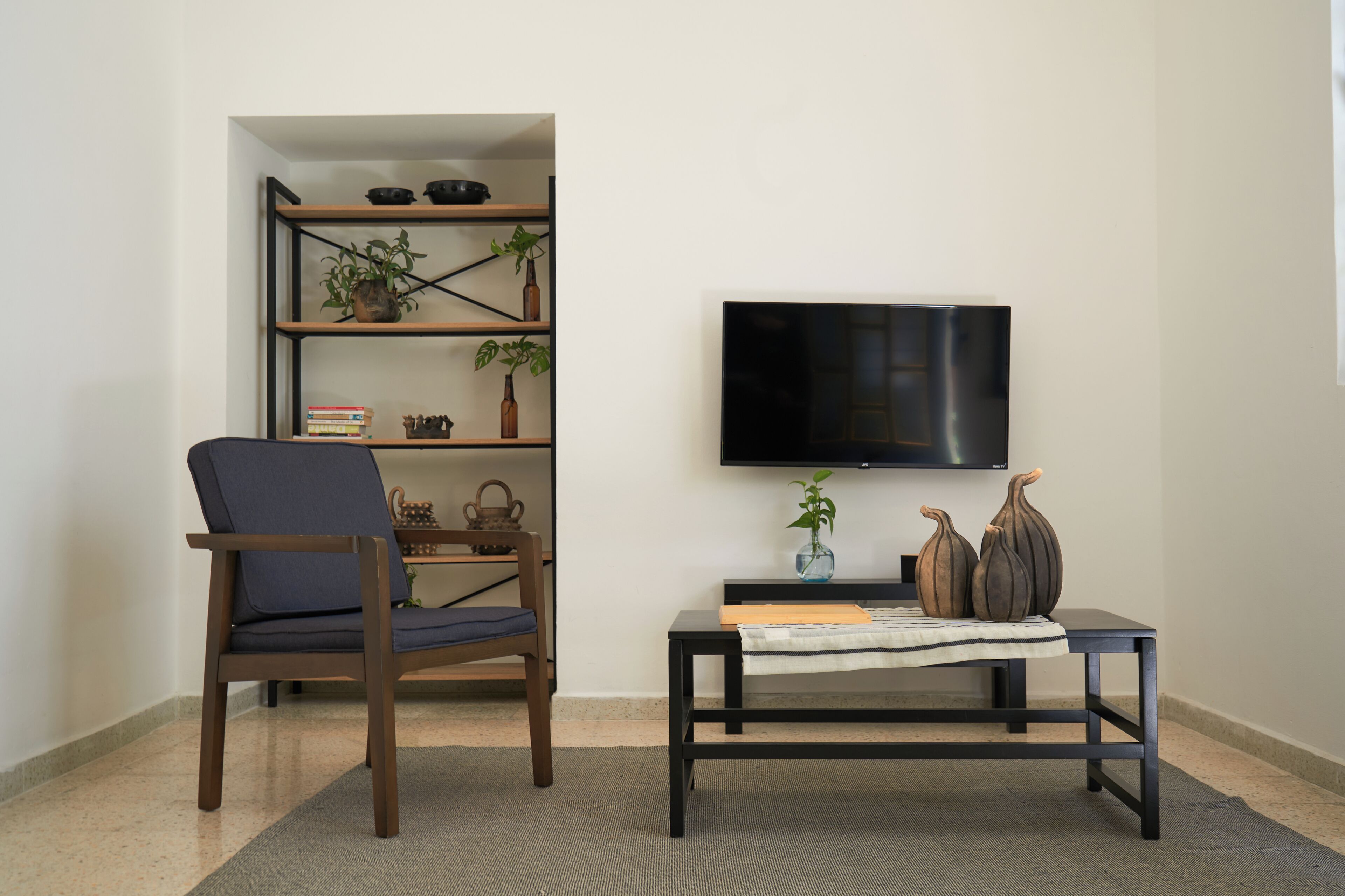 Una sala de estar con una silla de madera oscura, una mesita con un jarrón y libros, y un televisor en la pared.