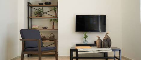 Una sala de estar con una silla de madera oscura, una mesita con un jarrón y libros, y un televisor en la pared.