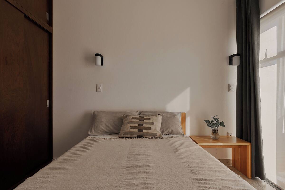 A neatly made bed with a patterned pillow, a bedside table with a plant, and a window with curtains.