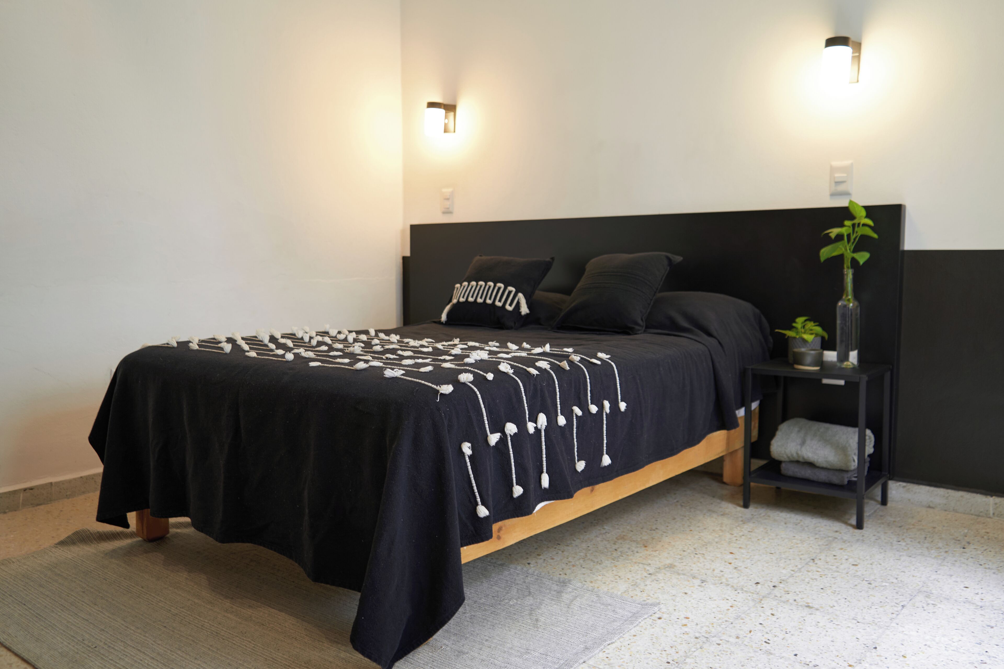 Un dormitorio moderno con una colcha negra, dos almohadas, una mesita de noche con una planta y luces fijadas a la pared.