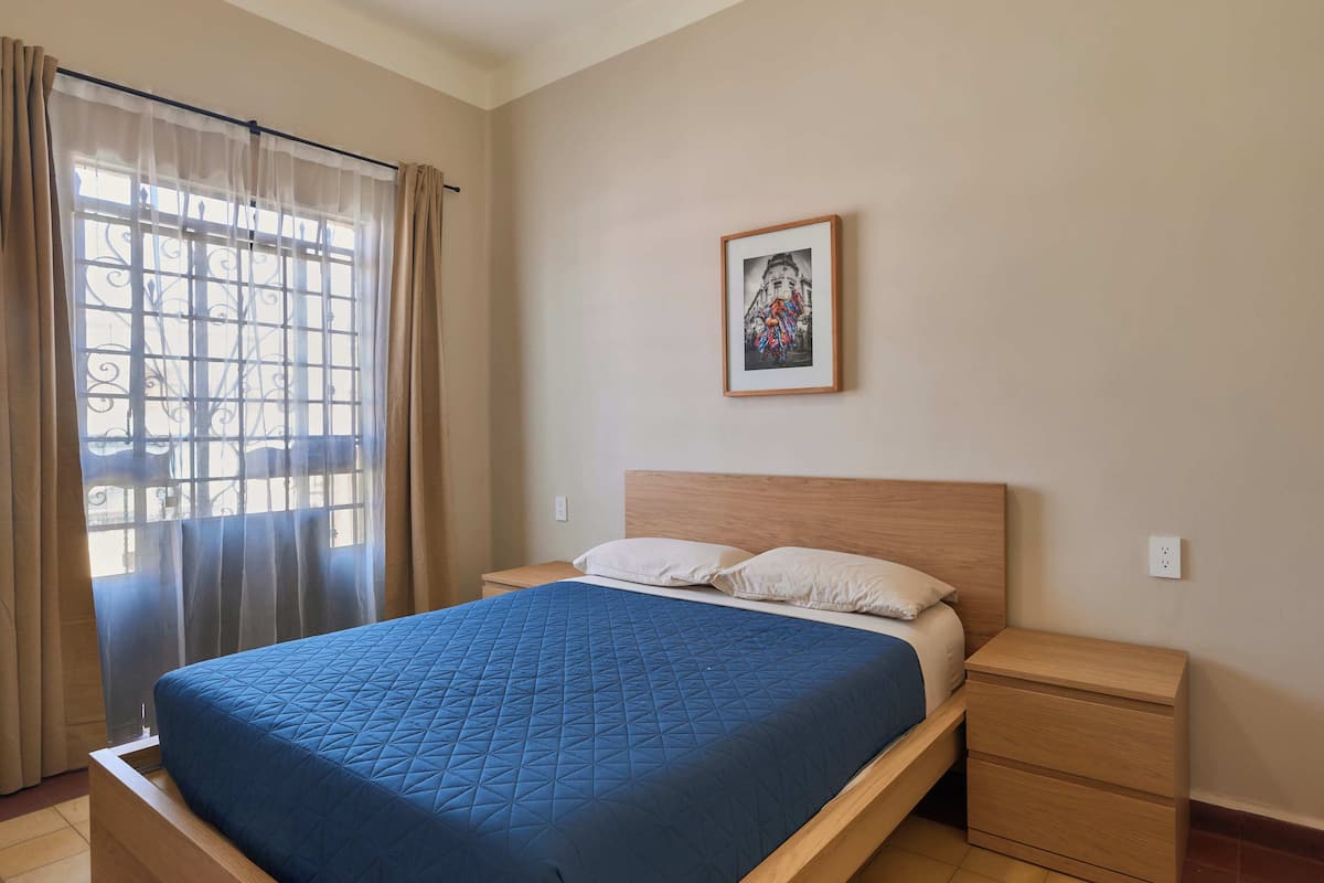 A bedroom with a bed, a nightstand, a wall-mounted picture, and a window with curtains.