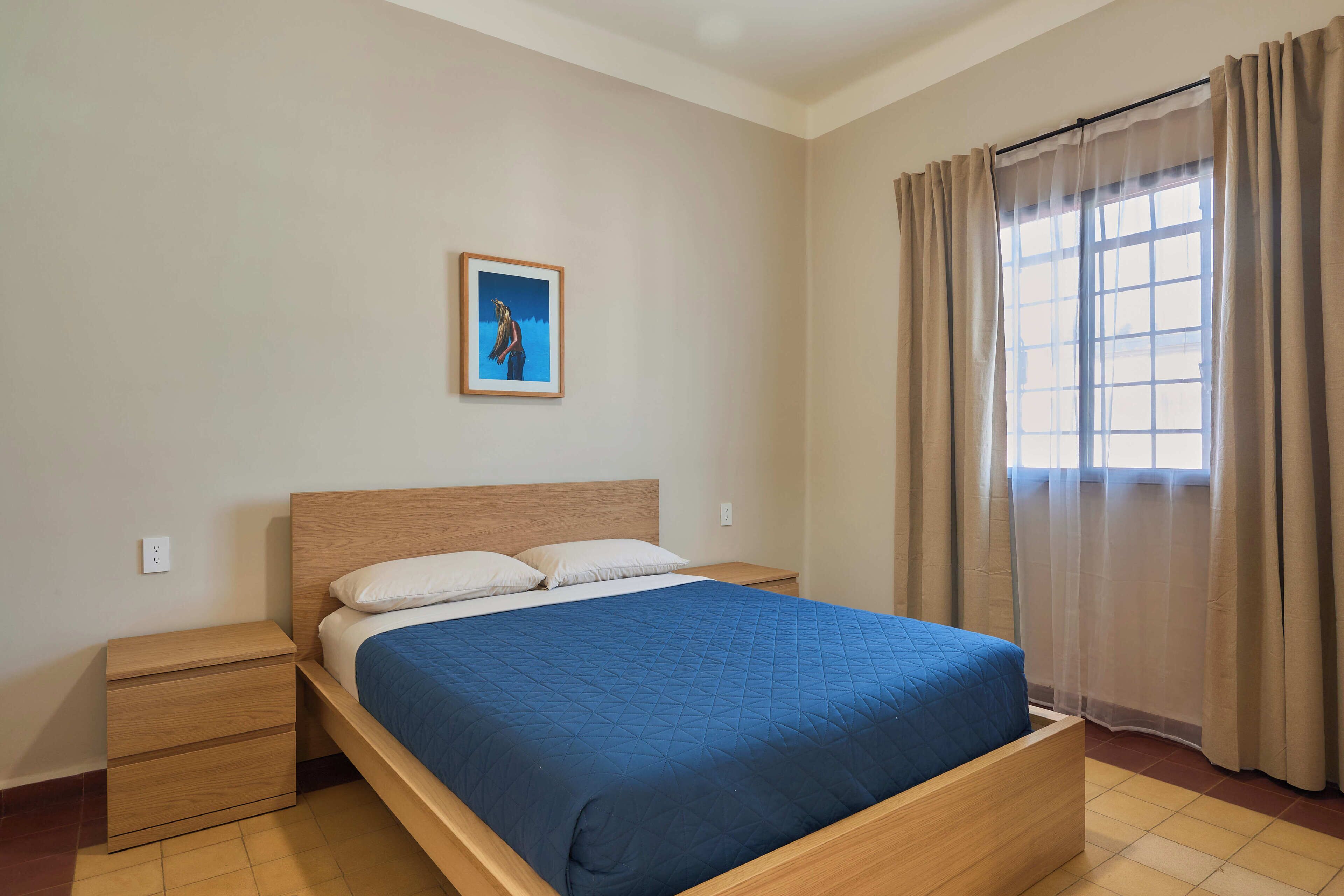 Een slaapkamer met een bed, nachtkastje, een aan de muur hangend schilderij en een raam met gordijnen.