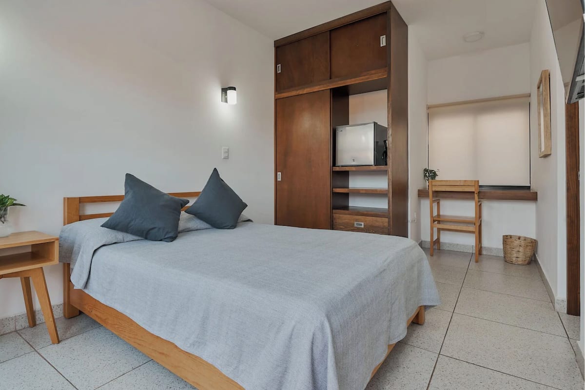 A bedroom with a bed, wooden wardrobe, desk, chair, and a small plant.