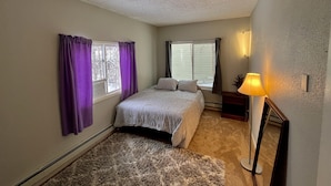 A bedroom with a bed, two windows with purple curtains, a nightstand, a lamp, and a mirror. - Scenic Alaskan cabin in the mountains (Anchorage)