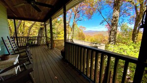 Um deck de madeira com guarda-corpo, ventilador de teto e vista para árvores e colinas.