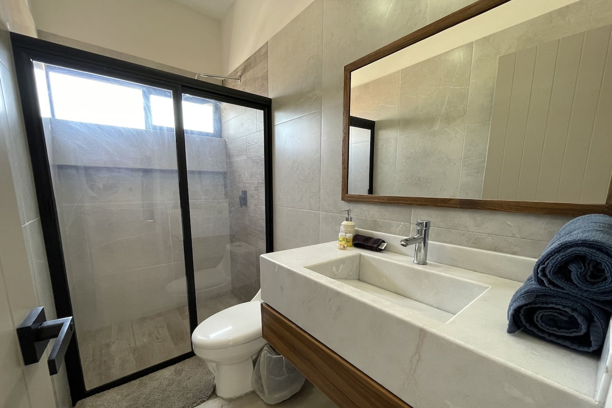 A modern bathroom with a glass-enclosed shower, a white toilet, a marble sink with a wooden base, a mirror above the sink, and a towel on the right side.