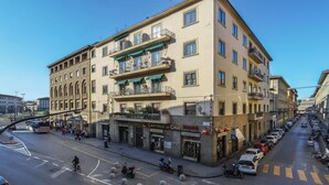 A multi-story building with shops on the ground floor, a clear sky, and a street with parked cars and a bus. - Santa Maria Novella Bright and Modern Apartment (Florence)