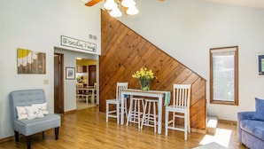 A living room with a wooden staircase, a dining area with white chairs, a blue sofa, and a ceiling fan. - Driftwood: Downtown Saugatuck Home, Walk to Shops & Restaurants, Dog-Friendly (Saugatuck)
