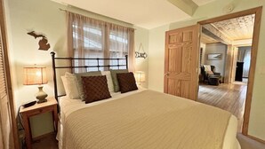 A bedroom with a bed, bedside lamps, a wooden door, and a view into another room. - Hillcrest: Downtown, Fire Pit, Fenced in Yard, Pet Friendly (Saugatuck)