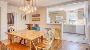 A dining area with a wooden table and chairs, a kitchen with white cabinets, and a hanging light fixture.
