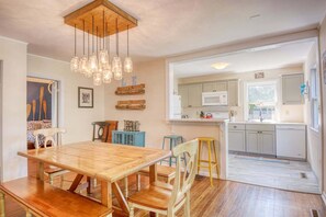 A dining area with a wooden table and chairs, a kitchen with white cabinets, and a hanging light fixture. - Lake Effect: Downtown Saugatuck, Pet-Friendly, Walk to Shops & Restaurants (Saugatuck)