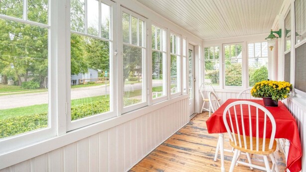 A covered porch with large windows, a table with a red tablecloth, and a chair. - Malabar: Downtown Saugatuck Cottage, Fire Pit, Pet Friendly, Sun Room, Porch (Saugatuck)