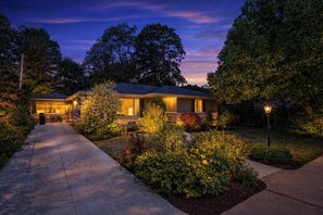 A two-story house with a covered patio and a well-lit garden at dusk.