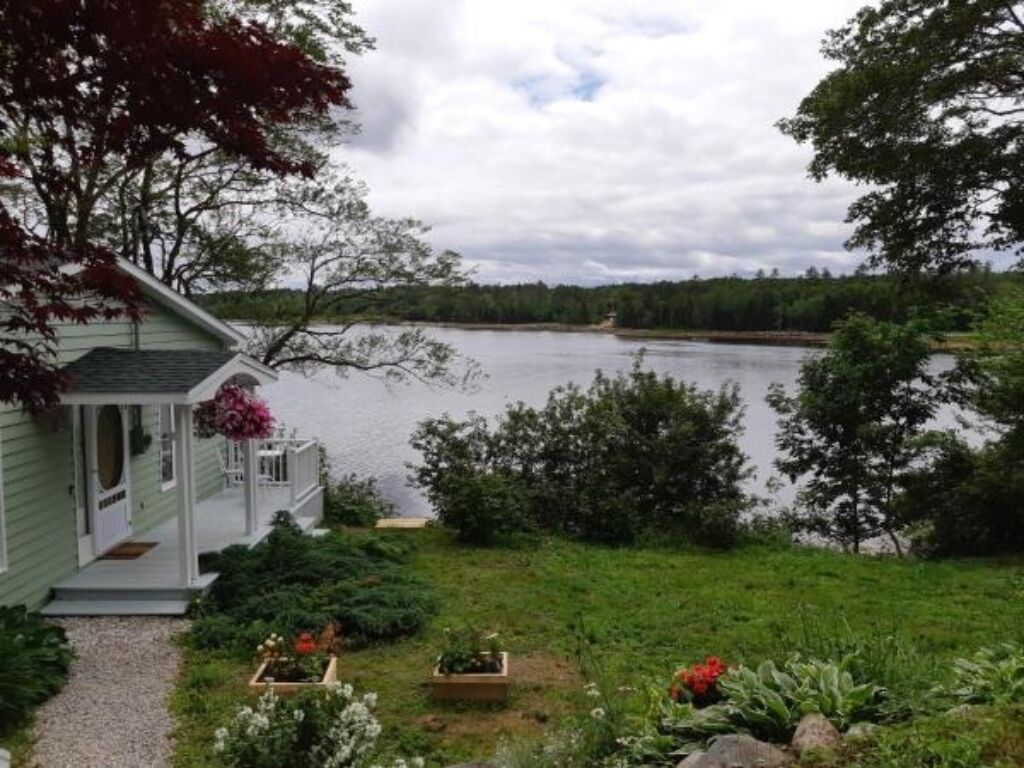 A house with a porch overlooking a lake, surrounded by greenery and trees.
