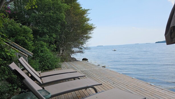 A wooden deck with lounge chairs and a table, overlooking a calm body of water with a distant island. - Oceanfront Garden Cottage - Gateway to Attractions & Towns. Private Cove Mooring (Chester)