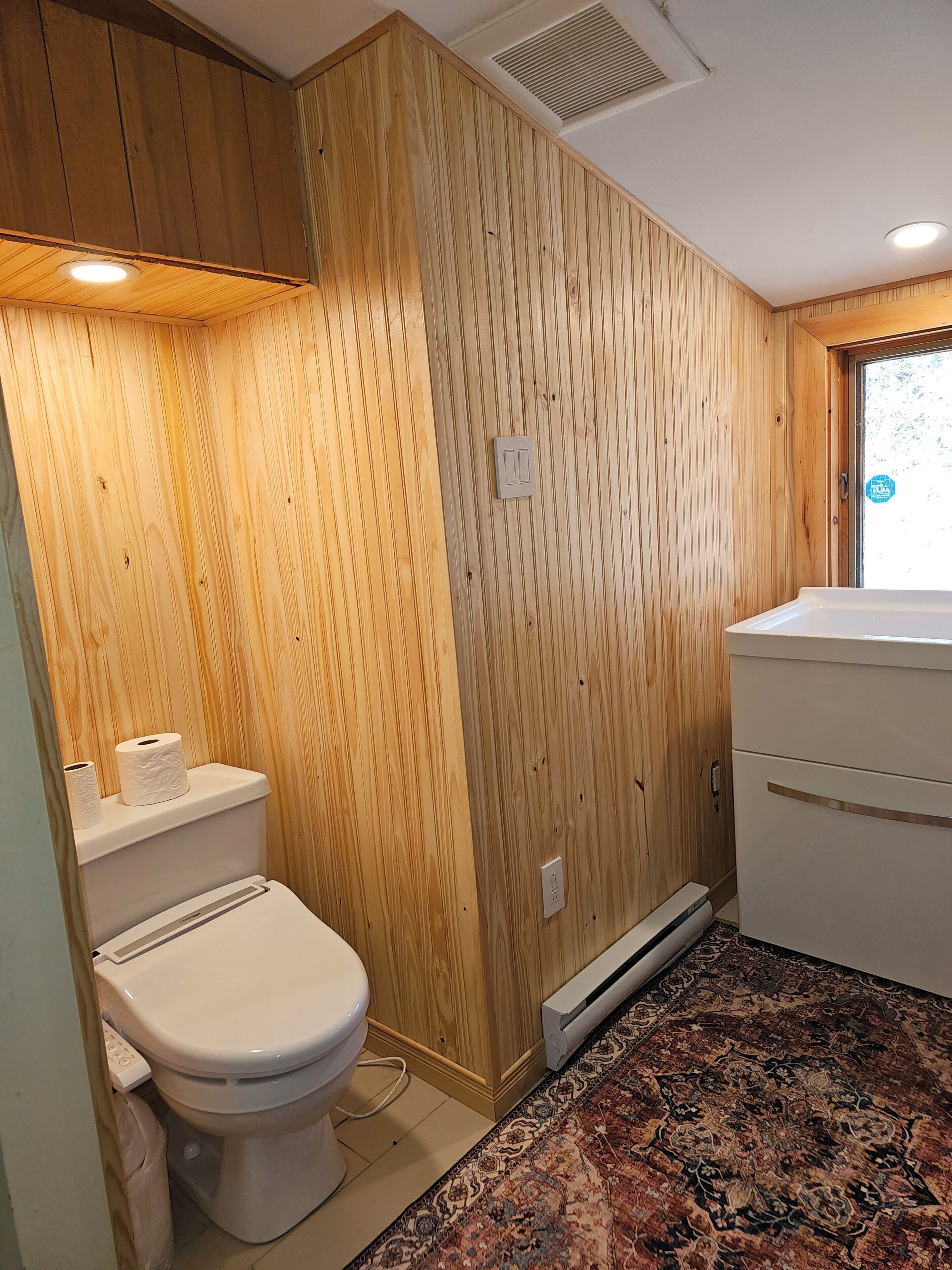 Un baño pequeño con paneles de madera, un inodoro, un radiador y una alfombra estampada.