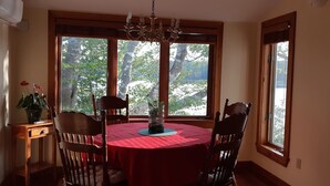 A dining room with a round table covered in a red tablecloth, surrounded by wooden chairs. There is a chandelier above the table, and the room has large windows with a view of trees and mountains outside. - Oceanfront Garden Cottage - Gateway to Attractions & Towns. Private Cove Mooring (Chester)