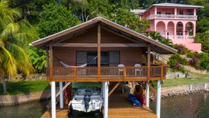 A wooden house with a balcony, a boat docked underneath, and a pink building in the background.
