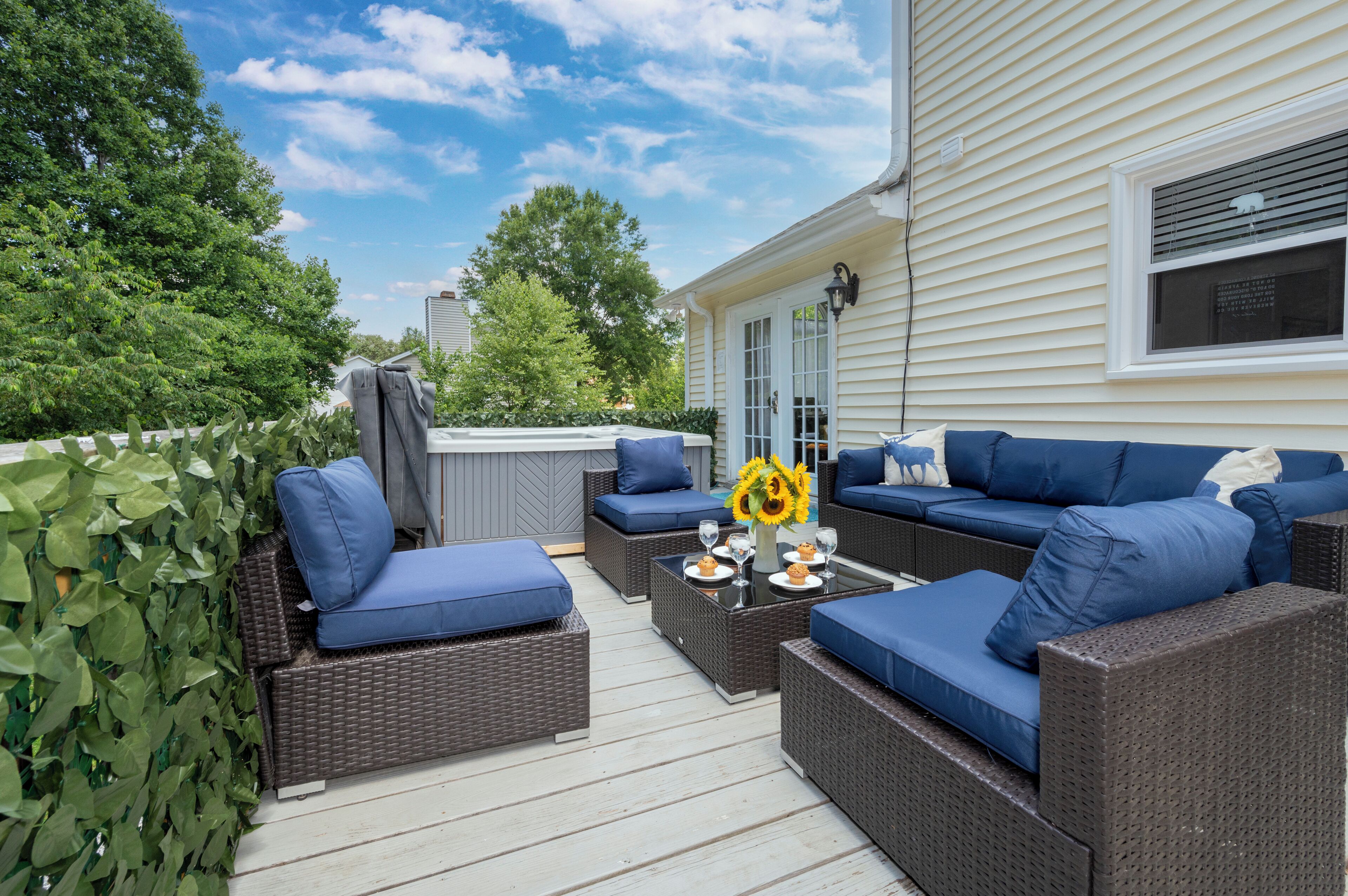 Une terrasse extérieure avec des meubles en osier et des coussins bleus, une table en verre surmontée d’un vase rempli de tournesols, et une vue sur une maison en arrière-plan.