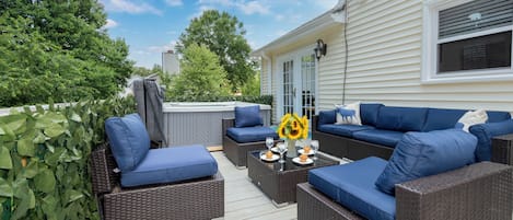 Une terrasse extérieure avec des meubles en osier et des coussins bleus, une table en verre surmontée d’un vase rempli de tournesols, et une vue sur une maison en arrière-plan.
