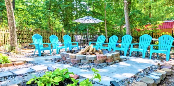An outdoor seating area with multiple Adirondack chairs arranged around a fire pit, surrounded by a wooden fence and trees.