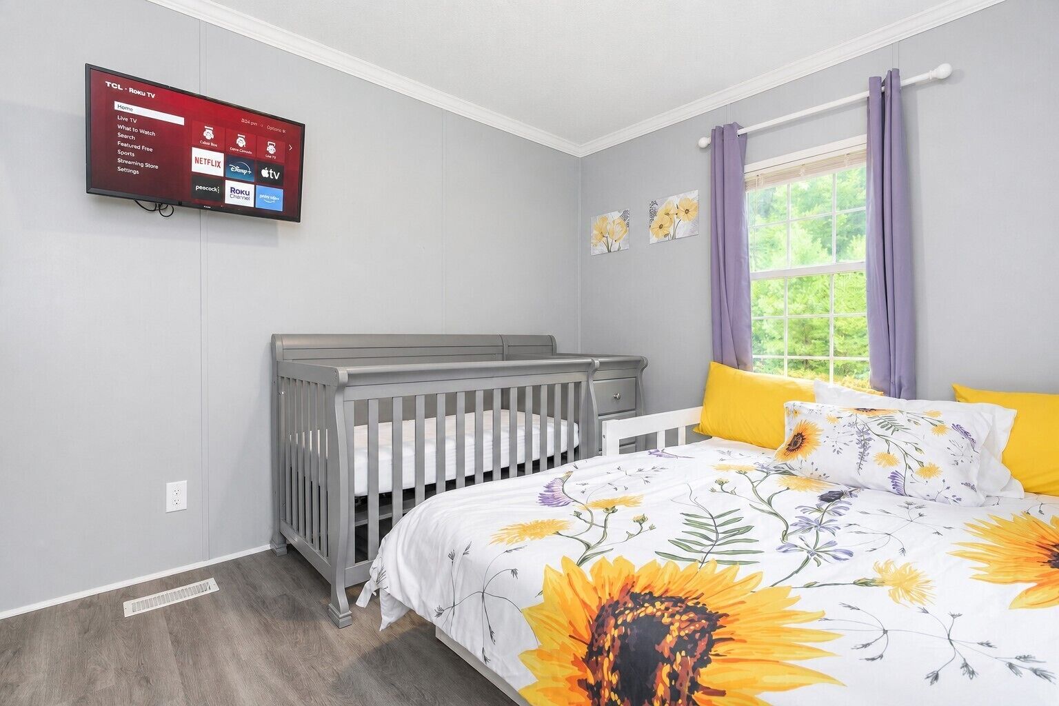 Ein Schlafzimmer mit einem Kinderbett, einem Bett mit Blumenmuster auf der Bettwäsche, einem an der Wand montierten Fernseher, der Streaming-Dienste anzeigt, und einem Fenster mit Vorhängen.