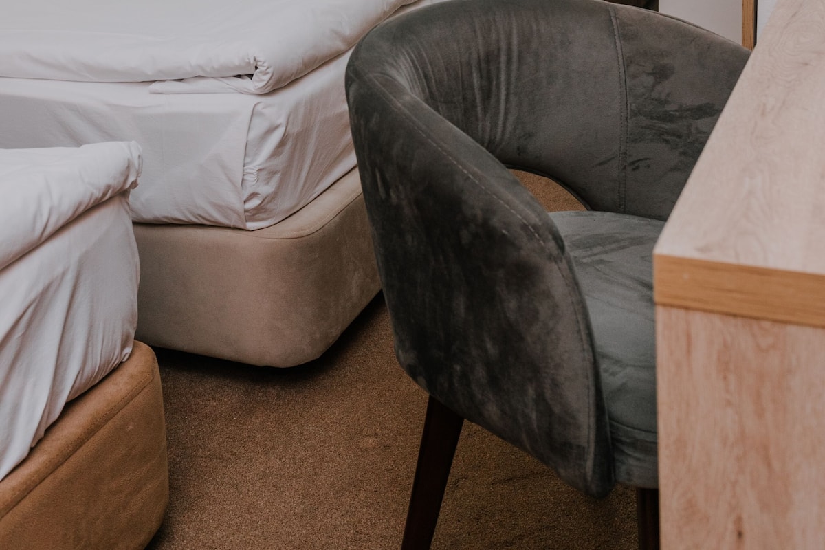 Ein Hotelzimmer mit zwei Betten, einem grau gepolsterten Sessel und einem Holztisch.