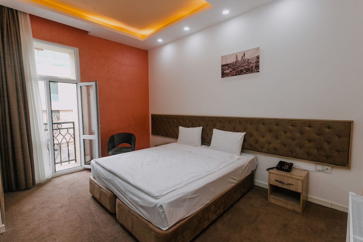 Ein Hotelzimmer mit einem großen Bett, einem Nachttisch mit Telefon, einem Fenster mit Vorhängen und einem Stadtbild an der Wand.