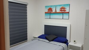 Een slaapkamer met een bed, een nachtkastje, een aan de muur hangend schilderij, een raam met jaloezieën en een kleine potplant.