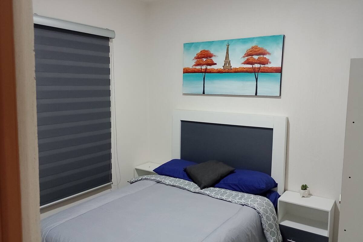 Een slaapkamer met een bed, een nachtkastje, een aan de muur hangend schilderij, een raam met jaloezieën en een kleine potplant.