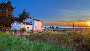 Una casa costera con un amplio balcón, rodeada de vegetación natural y vistas al océano al atardecer.