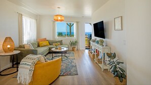 Una sala de estar con sofá, sillón, mesa de centro y televisor. Se ve un ventanal con vistas al exterior.
