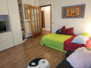 Um quarto com uma cama, uma televisão, um guarda-roupa e uma porta que dá para outro cômodo.