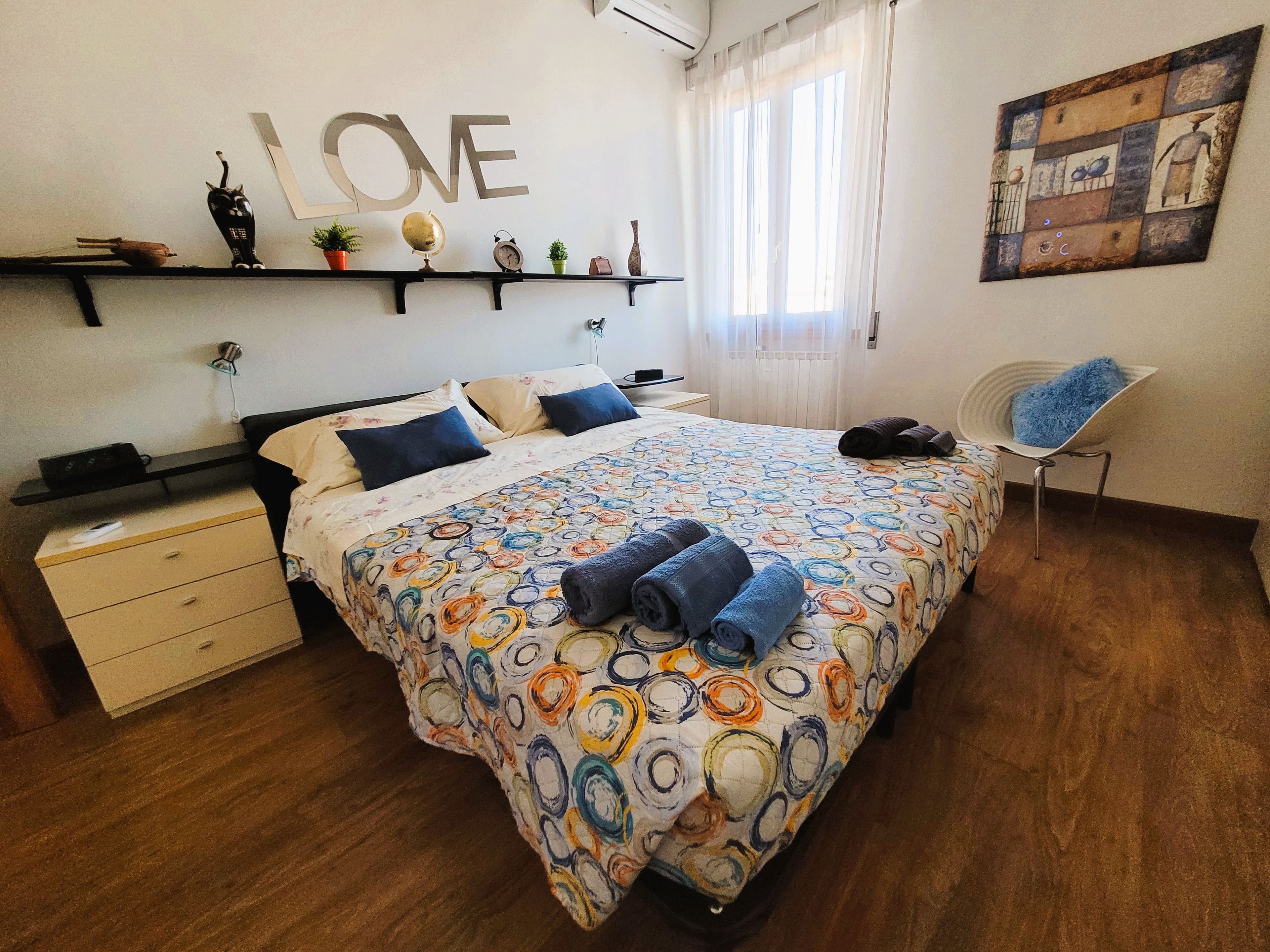 Ein Schlafzimmer mit einem Bett, einem Sessel, einem Regal mit Dekoration und einem Wandbild mit der Aufschrift 'LOVE', sowie einem Gemälde.