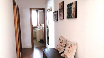 Un pasillo con piso de madera, un banco con cojines estampados, una lámpara de pared y cuadros enmarcados.