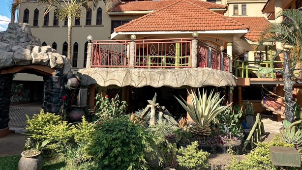 Un hotel con techo de tejas rojas, un balcón con barandilla de hierro forjado y un jardín con diversas plantas y una palmera.