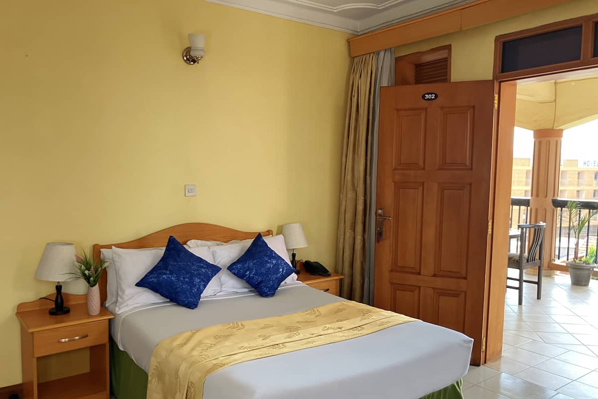 A hotel room with a bed, nightstands, a lamp, a potted plant, and a door leading to an outdoor area.