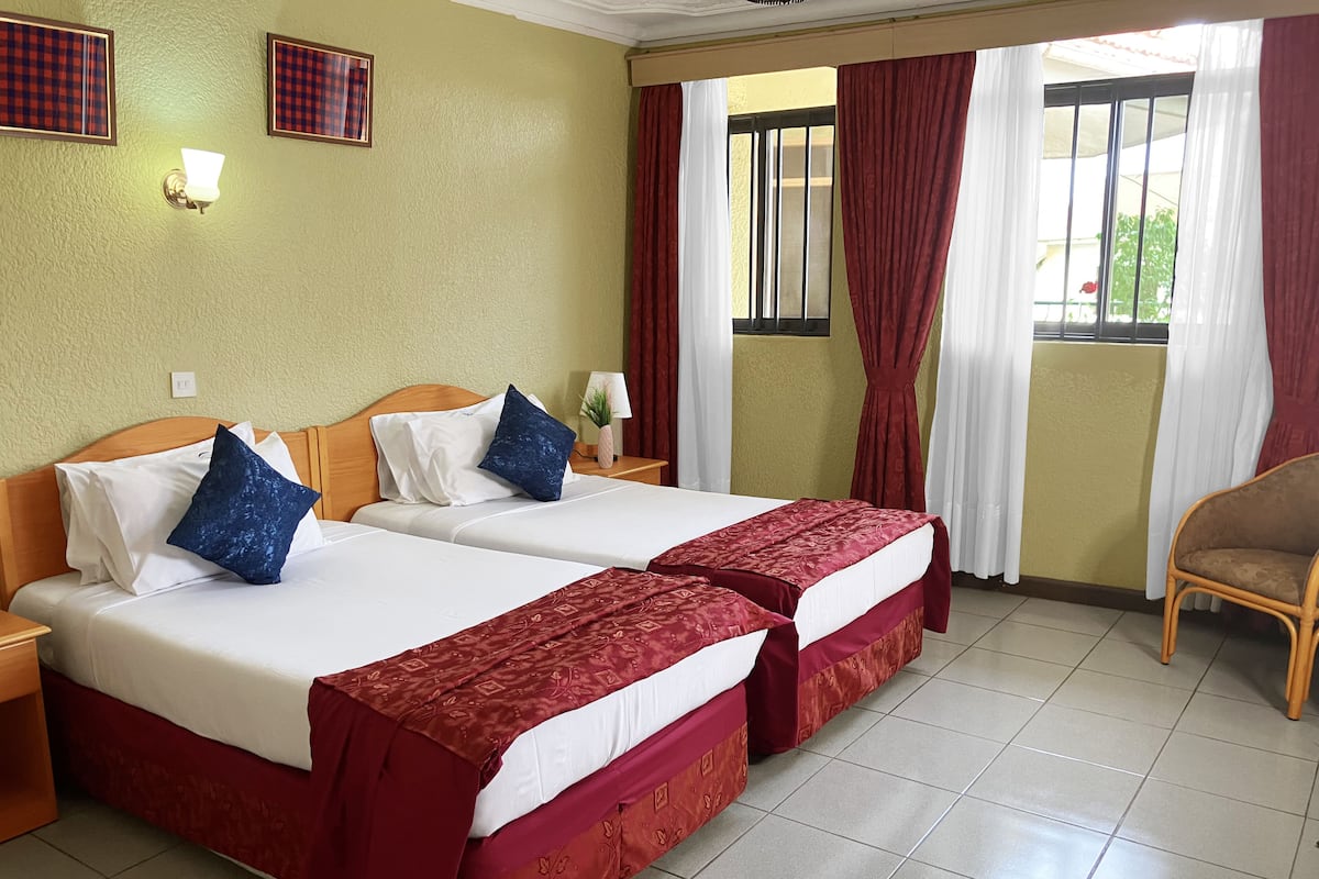A hotel room with two beds, a wooden headboard, a nightstand, a lamp, and a window with curtains.