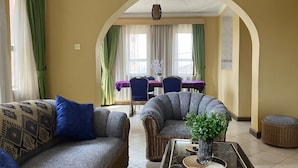 Una sala de estar con un sofá, un sillón y una mesa de centro con un florero. En el fondo, hay un comedor con una mesa y sillas.
