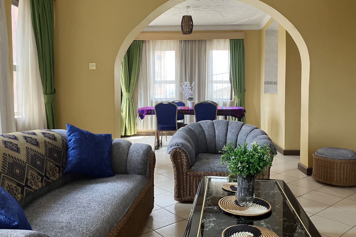 A living room with a sofa, armchair, and a coffee table with a vase. There is a dining area with a table and chairs in the background.