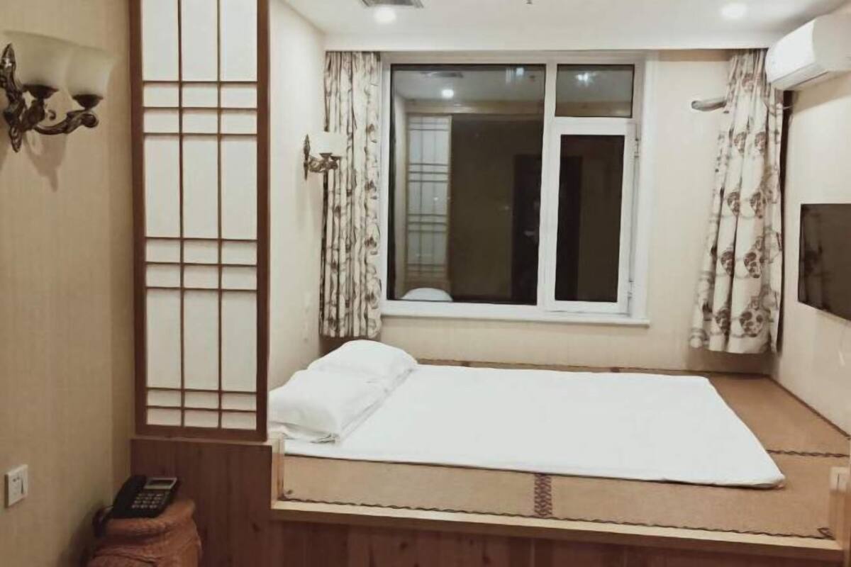 A small, clean room with a single bed, a window with curtains, and a wall-mounted TV.