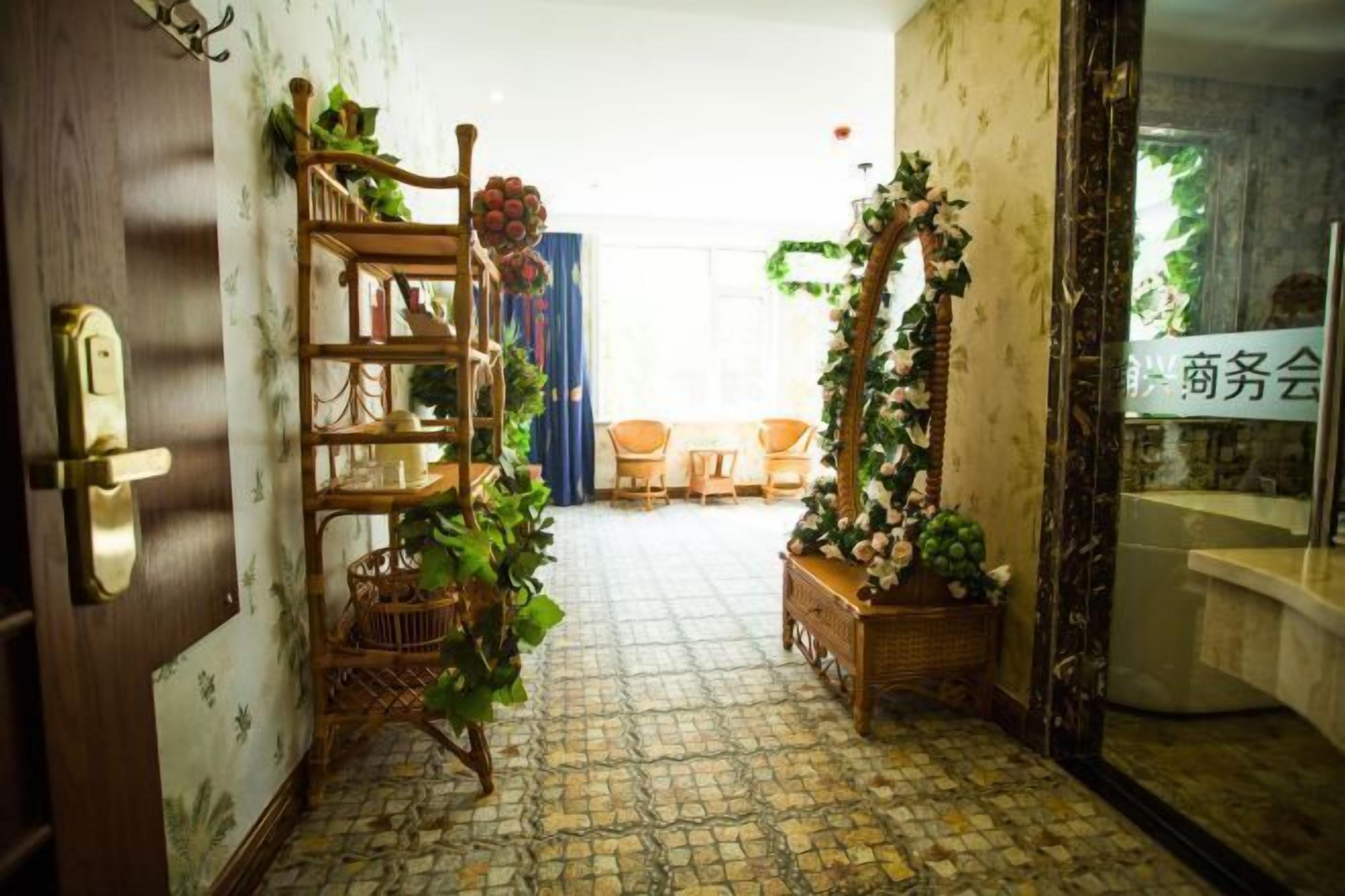 A hallway with a wooden door, a wicker shelf with plants, a tiled floor, and a view into a room with a table and chairs.