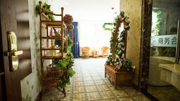 A hallway with a wooden door, a wicker shelf with plants, a tiled floor, and a view into a room with a table and chairs.