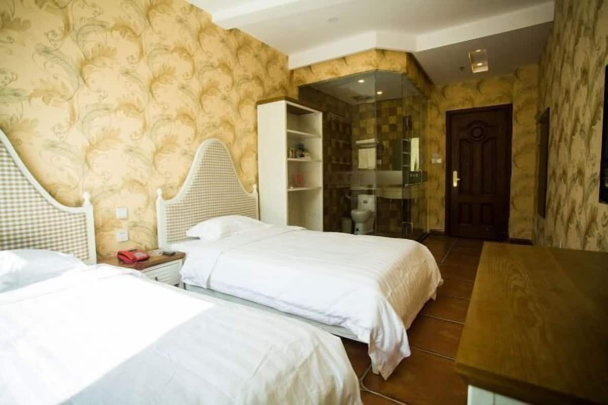 A hotel room with two beds, a wardrobe, a bathroom with a glass shower, and a wooden table.