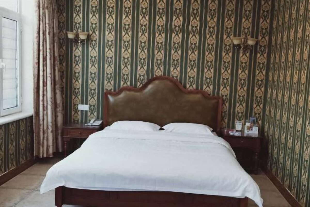 A neatly made bed with a wooden headboard in a room with patterned wallpaper and a window with curtains.