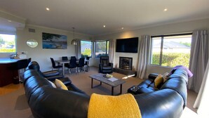 A spacious living area with a black leather sofa, a coffee table, and a fireplace. There is a dining area with chairs and a table, and a TV mounted on the wall.
